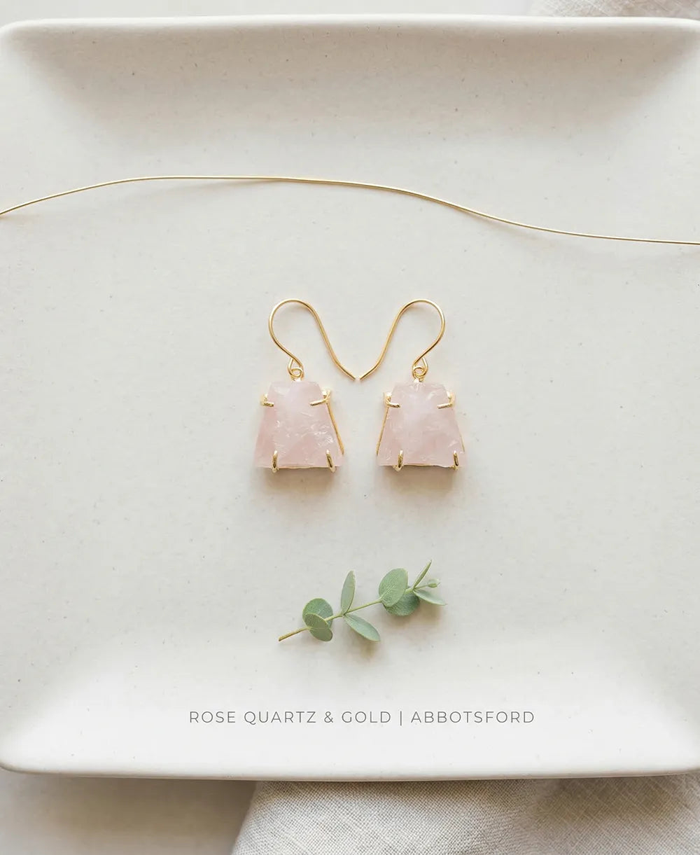 Rose quartz earrings with gold accents on a white plate with eucalyptus leaves.