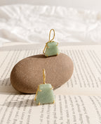 Two green stone earrings with gold wire frames on a beige stone and newspaper background.