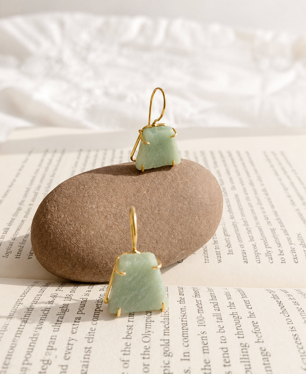 Two green stone earrings with gold wire frames on a beige stone and newspaper background.