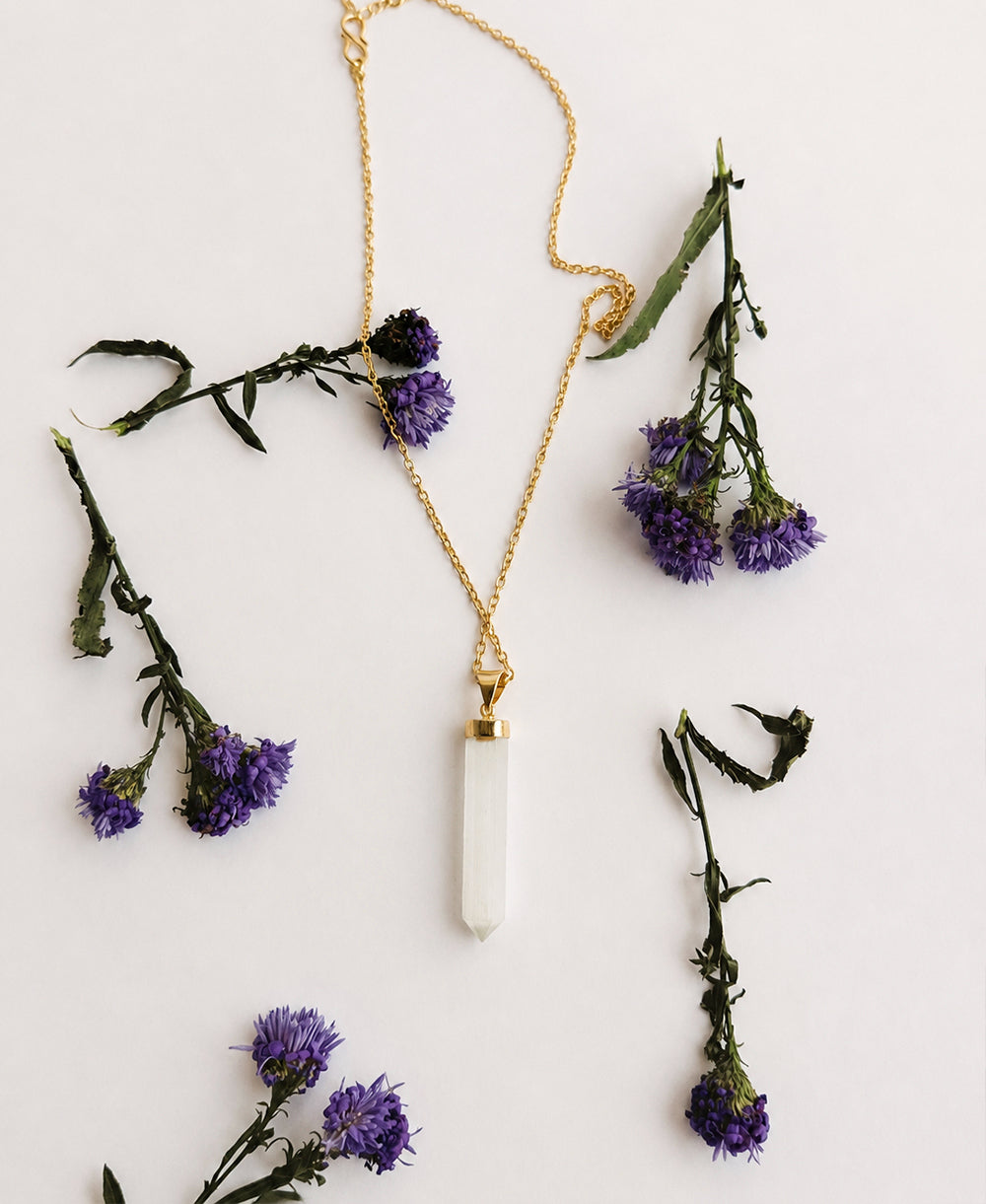 Gold necklace with a white pendant surrounded by purple flowers on a light background